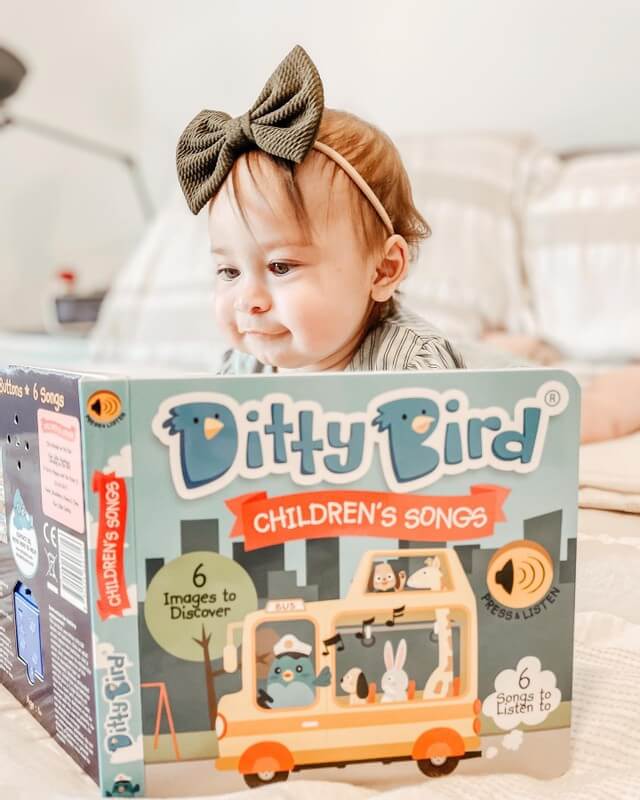 A baby with a large green bow headband sits on a bed, looking at the Books brand Children's Songs interactive sound book (USB-C rechargeable), which features colorful cartoon animals and classic children's songs on its cover.