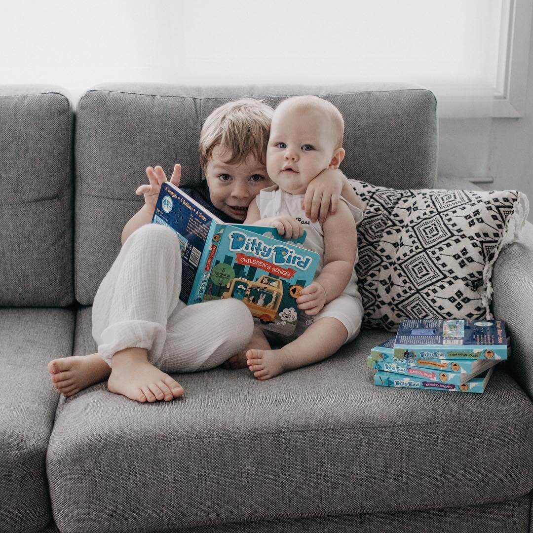 Two young children sit on a grey couch; the older hugs the younger while reading "Children's Songs" by Books, an interactive sound book that's USB-C rechargeable. Nearby are more books and a patterned cushion.