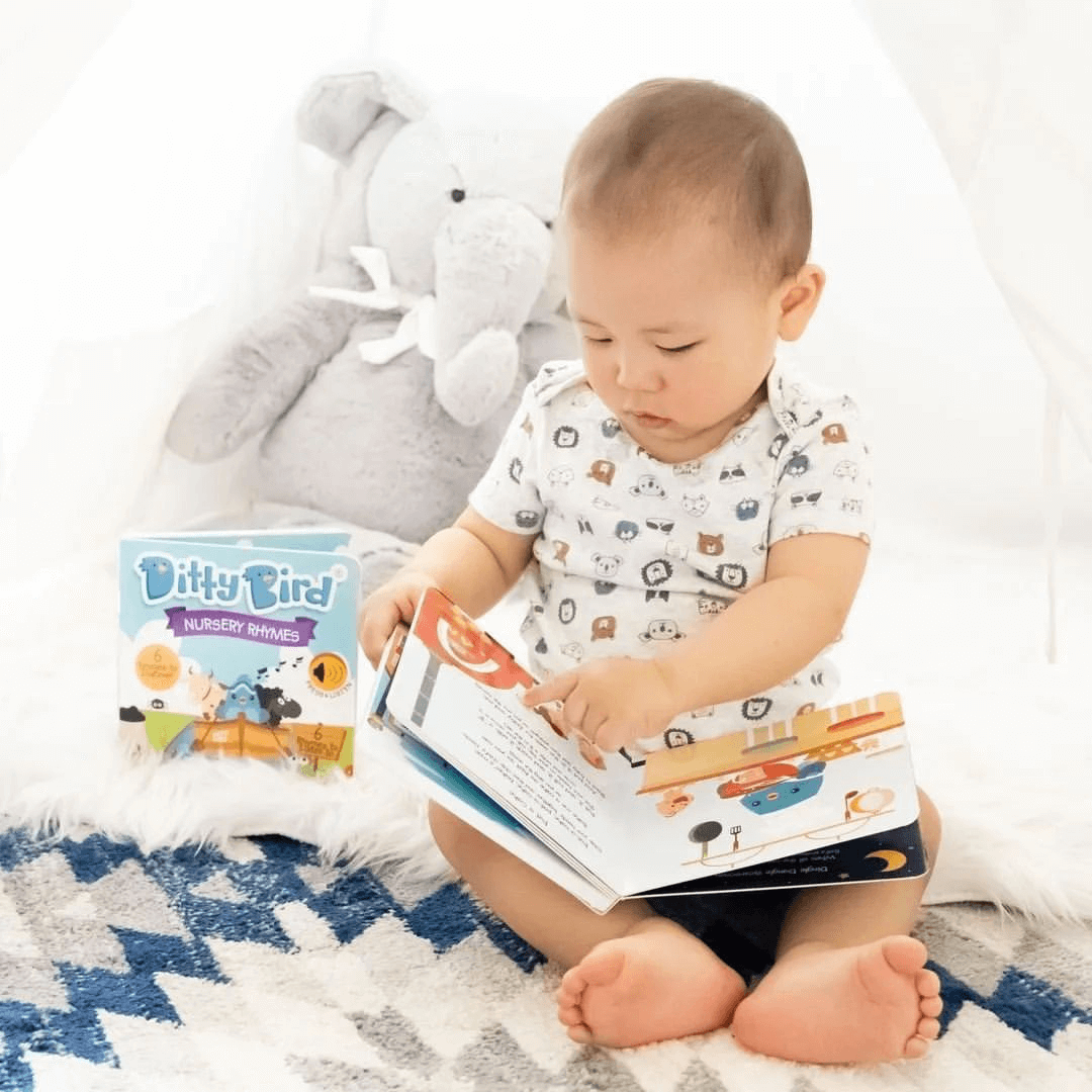 A baby sits on a blue and white patterned blanket, looking at a picture book. Nearby is a Ditty Bird Nursery Rhymes USB-C rechargeable sound book and a gray stuffed elephant.