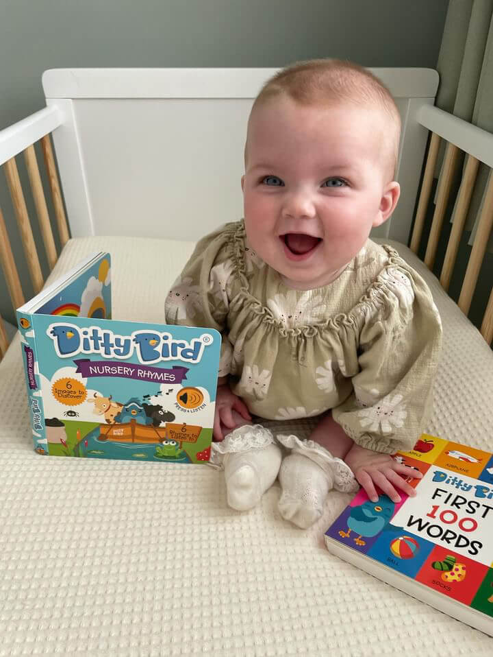 A smiling baby in a light green dress sits on a crib mattress, holding Ditty Bird’s FIRST 100 WORDS musical book, with the Ditty Bird Nursery Rhymes | USB-C rechargeable sound book open in front of them.