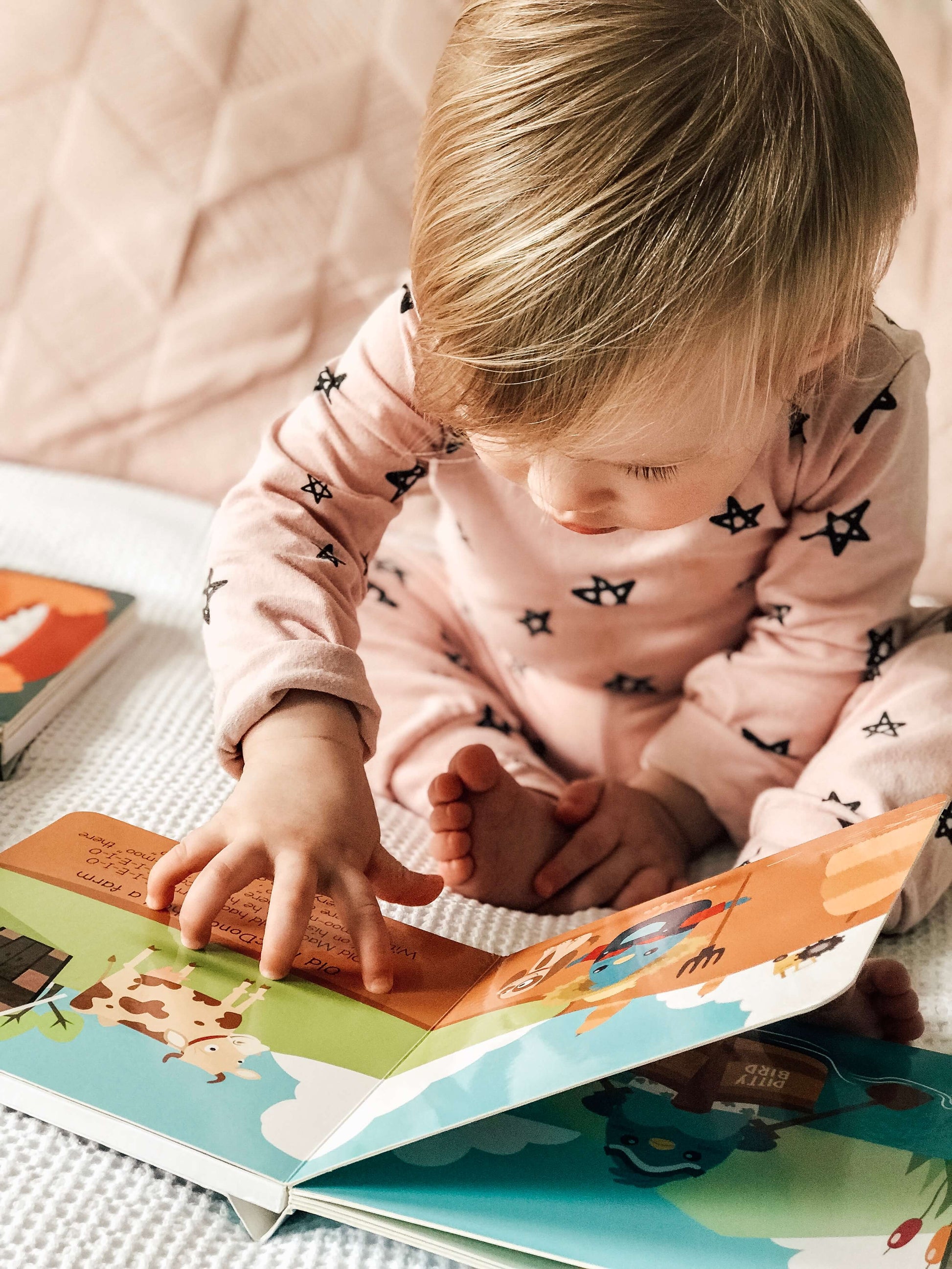 A toddler in pink pajamas with black stars sits on a bed, looking down and exploring the colorful Ditty Bird Nursery Rhymes musical book by Ditty Bird, USB-C rechargeable and filled with animal illustrations.
