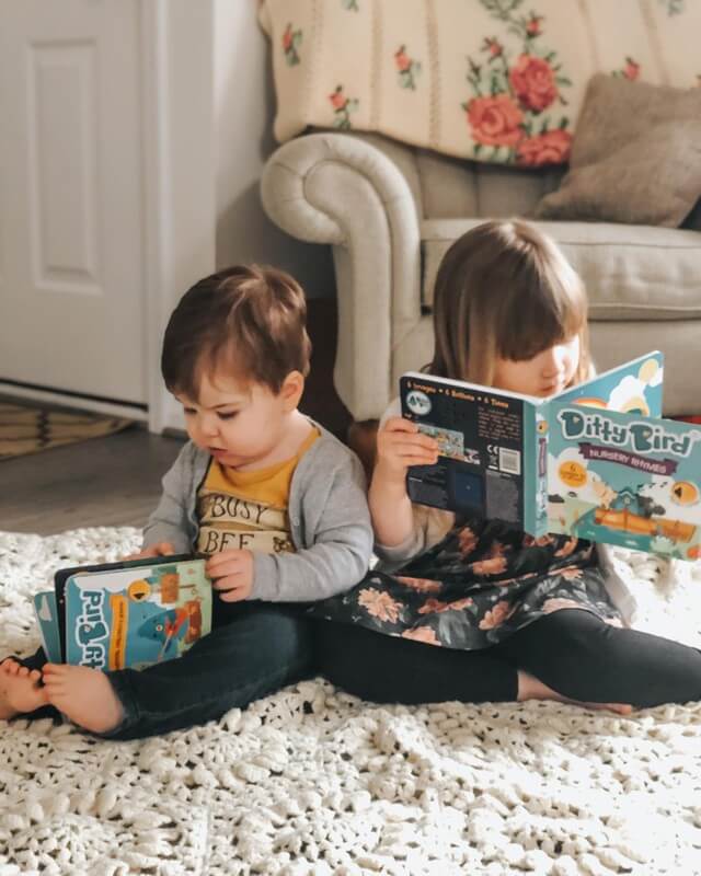 Two young children sit back-to-back on a cream rug, each enjoying a Ditty Bird Nursery Rhymes | USB-C rechargeable sound book. A couch with a floral blanket enhances the cozy indoor setting.