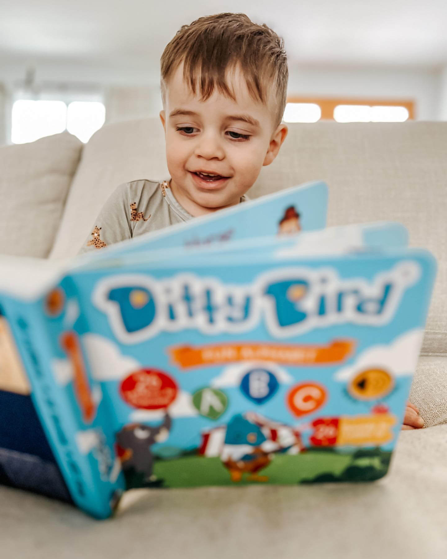 A young child with light brown hair smiles on a couch, engaging with the colorful Ditty Bird ABC Fun Alphabet | AAA model sound book, creating a cheerful early literacy scene in a bright living room.