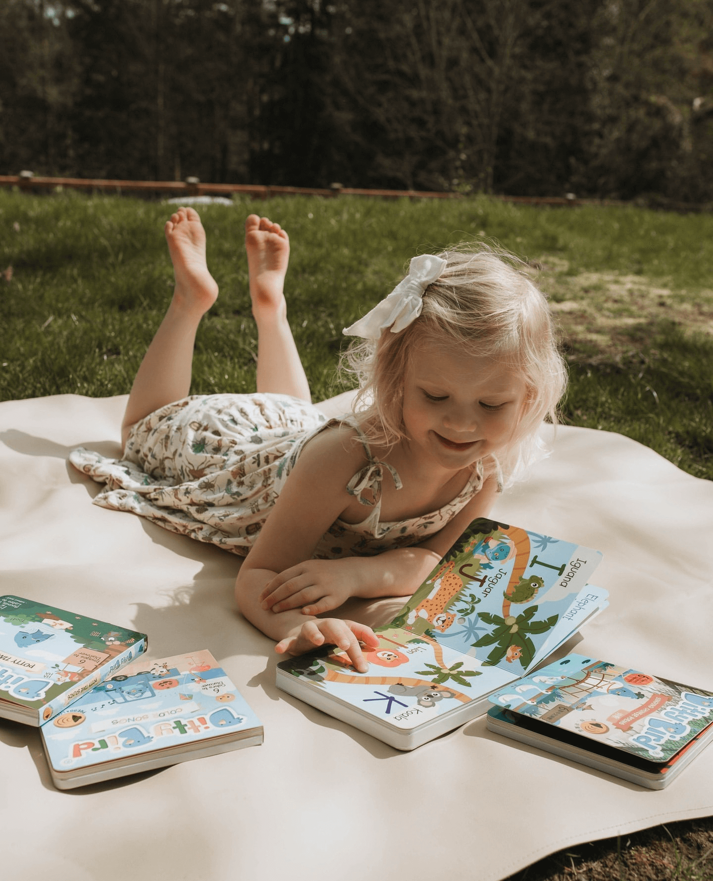 A young blonde girl with a white bow lies on a blanket outdoors, reading the Ditty Bird ABC Fun Alphabet | AAA model, with more books and interactive flashcards scattered nearby.