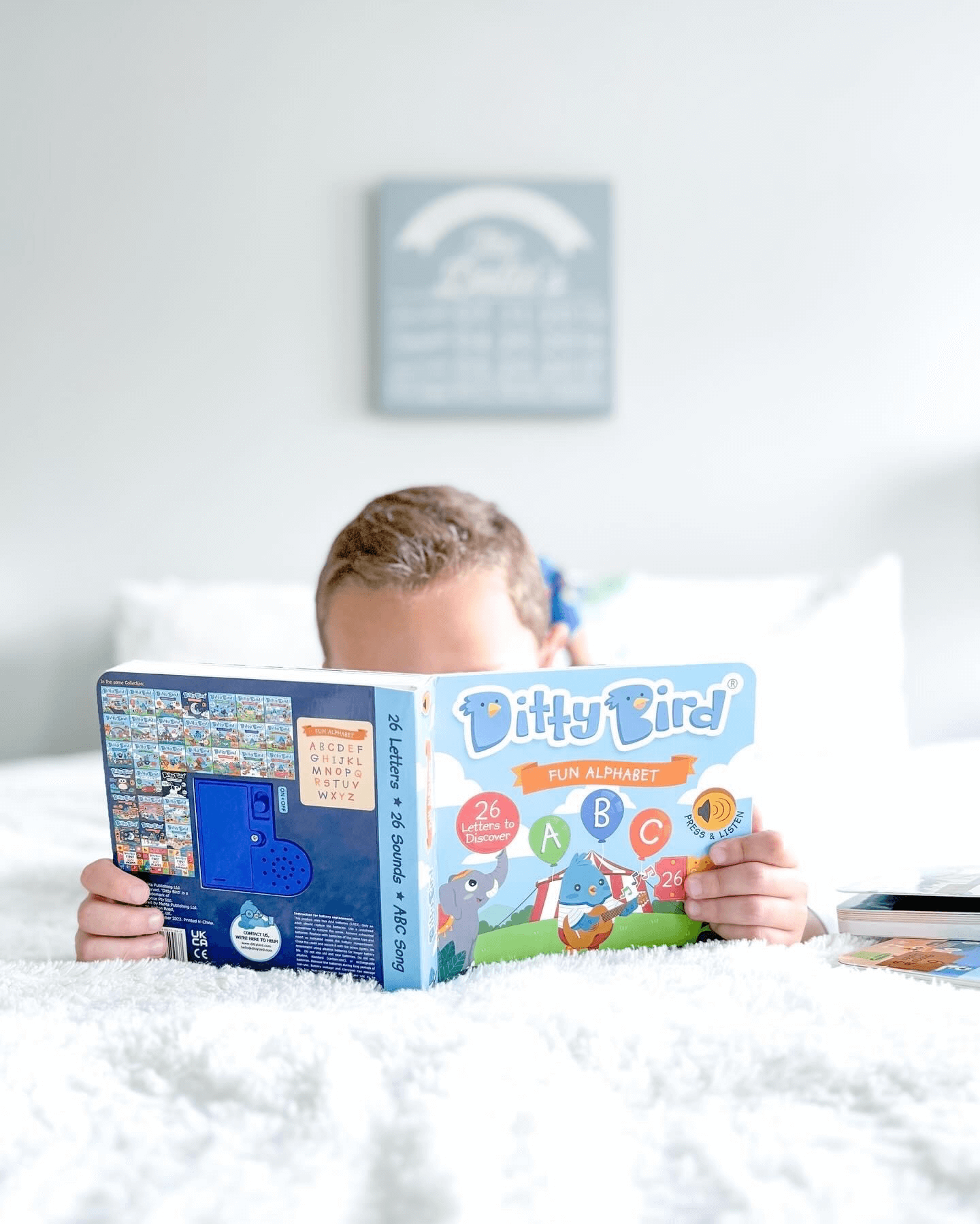 A child lies on a white bed, holding and reading the Ditty Bird ABC Fun Alphabet | AAA model book. The softly blurred background features a blue sign—perfect for fostering early literacy skills.
