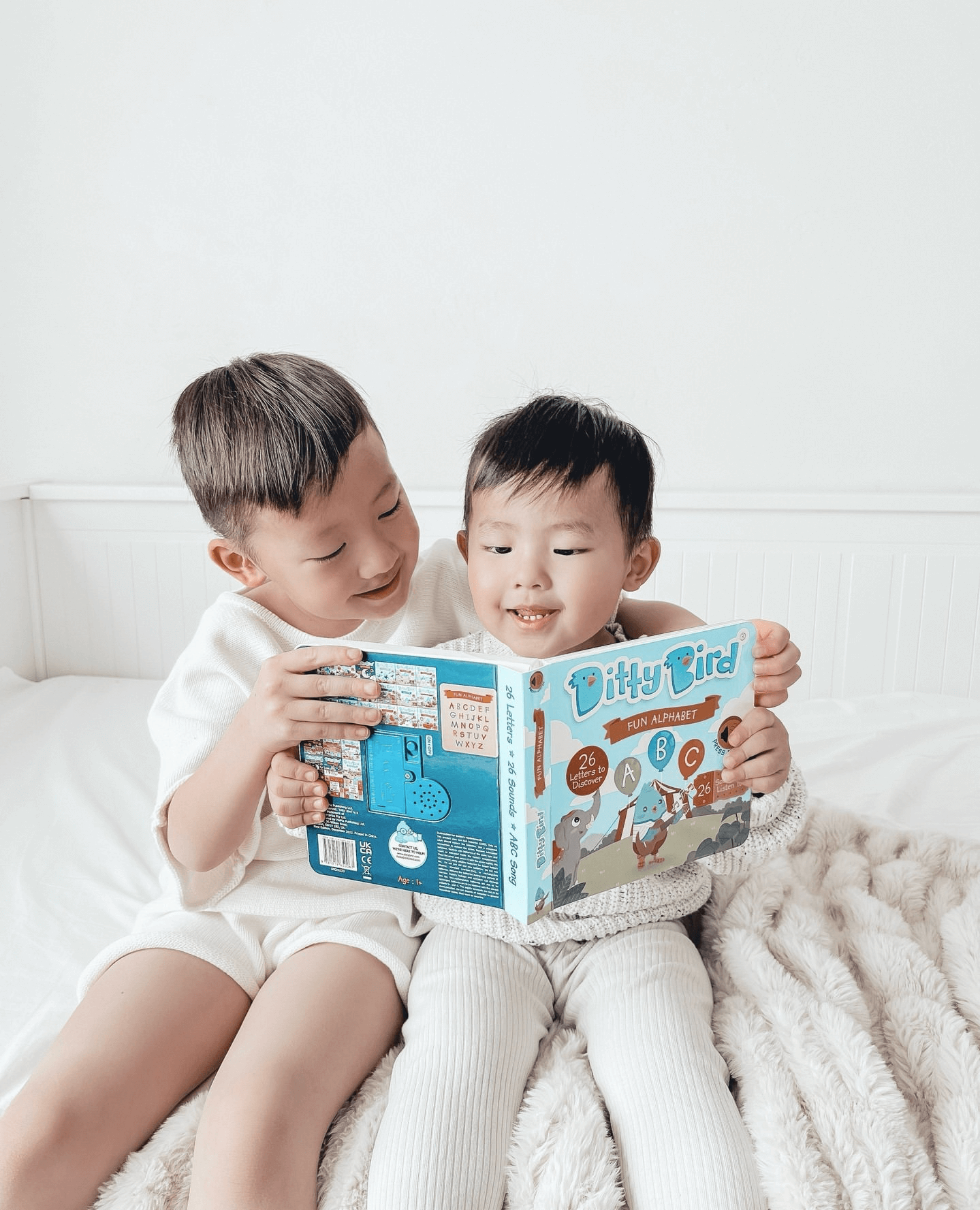 Two young children, smiling and engaged, sit on a bed reading the Ditty Bird ABC Fun Alphabet | AAA model book, enjoying early literacy together on a soft blanket.
