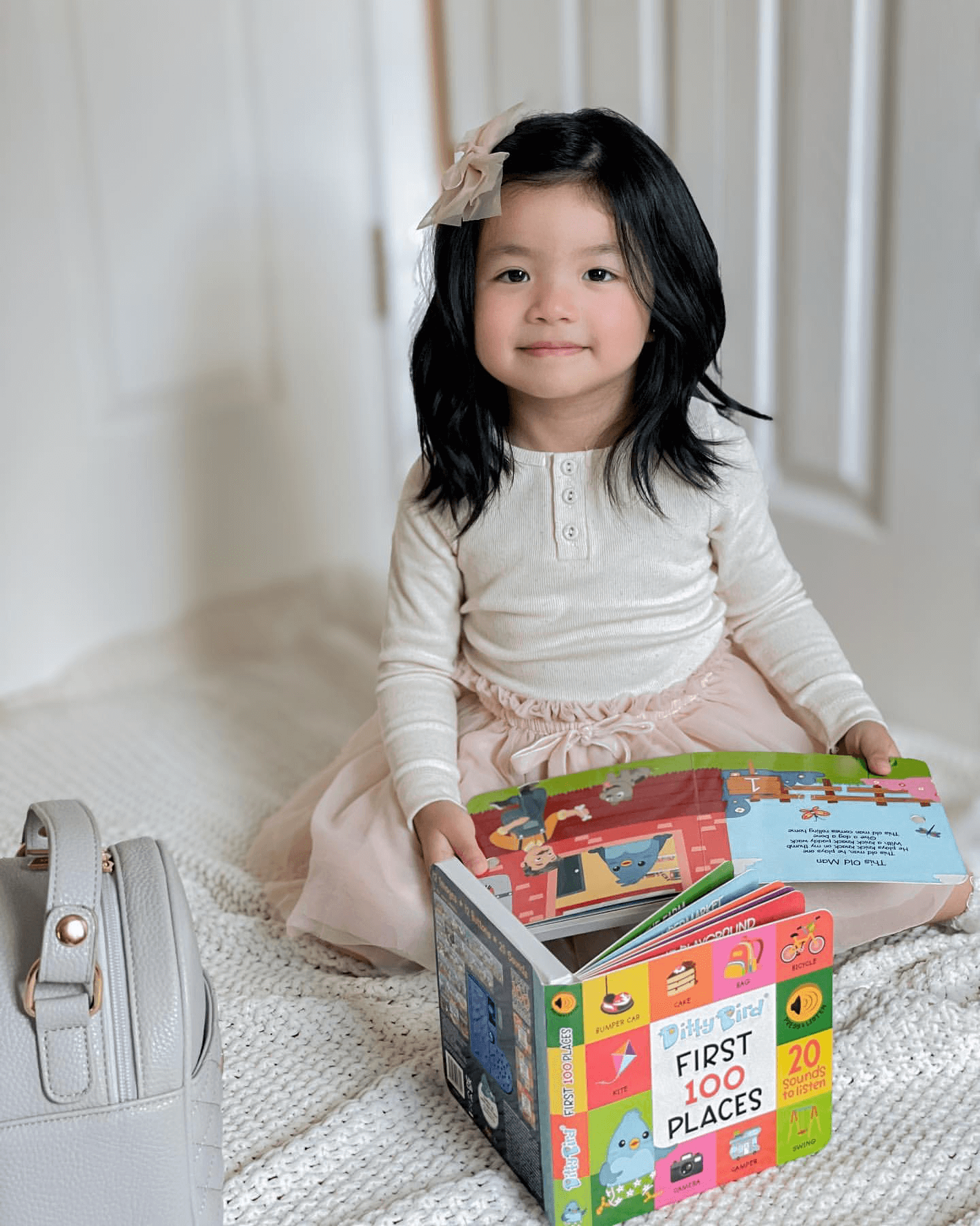 A young girl with long dark hair in a pale pink skirt and white top sits on a white blanket holding an open Ditty Bird Learning Songs interactive children’s book (USB-C rechargeable) next to a light gray handbag.