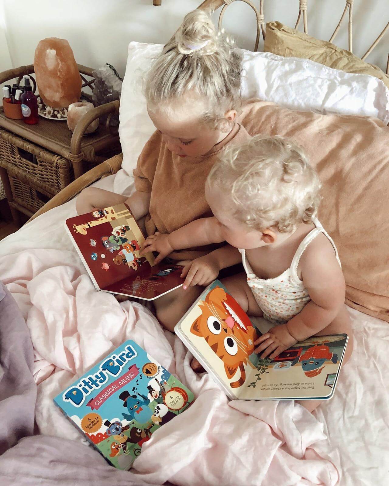Two blonde-haired children sit on a bed with cozy decor, sharing colorful picture books. A Ditty Bird Learning Songs Sound Book (USB-C rechargeable) is visible beside them. Soft lighting adds warmth to the scene.