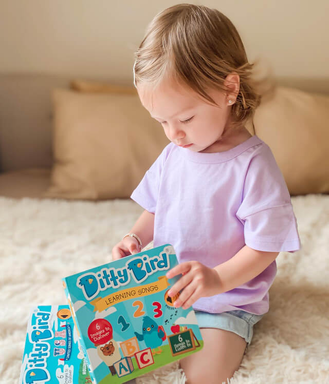 A light brown-haired toddler in a lavender shirt sits on a bed, focused and curious as they explore the Ditty Bird Learning Songs book—an interactive, USB-C rechargeable children’s book filled with colorful songs.