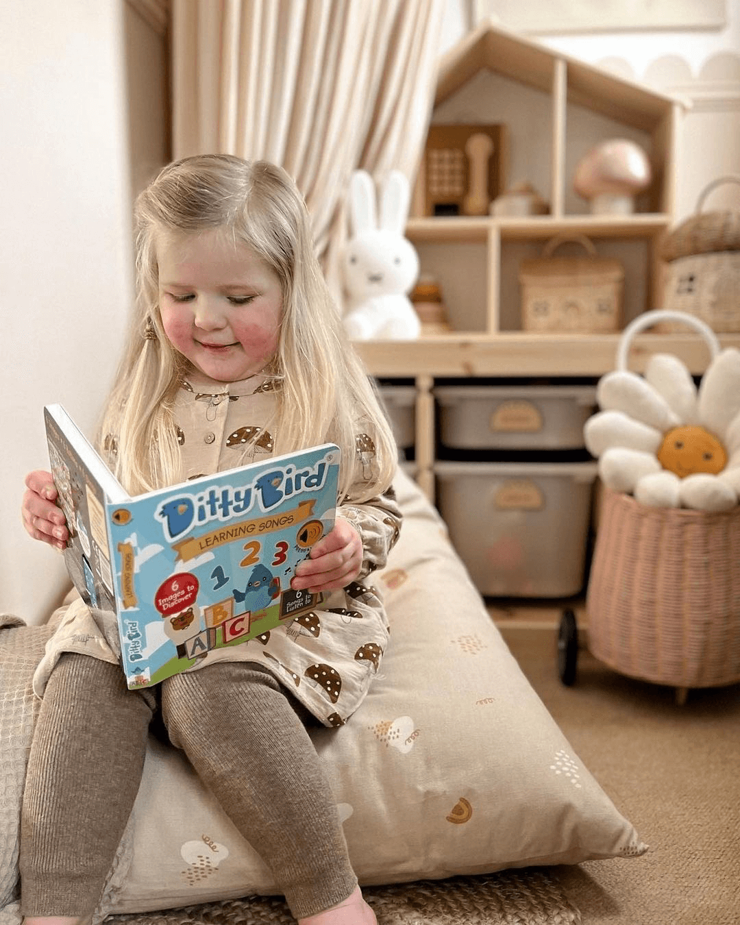 A young girl with long blonde hair sits on a beige cushion in a cozy playroom, reading the Ditty Bird Learning Songs interactive children's sound book (USB-C rechargeable), surrounded by toys, baskets, and a plush flower pillow.