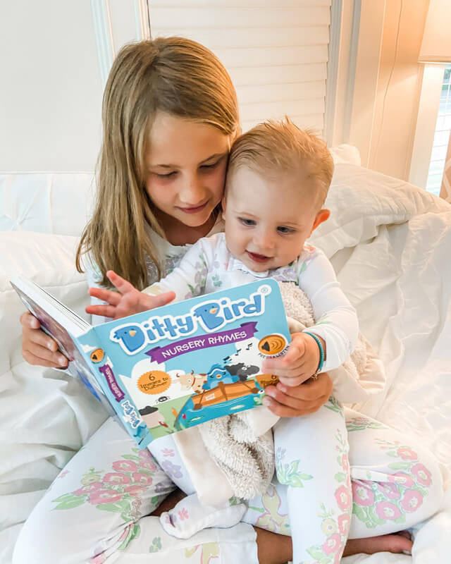 Two young children in pajamas sit on a bed, smiling as they read the Ditty Bird "Bedtime Songs" sound book together. The older child holds the book while the younger looks excitedly at the pages, enjoying interactive songs before bedtime.