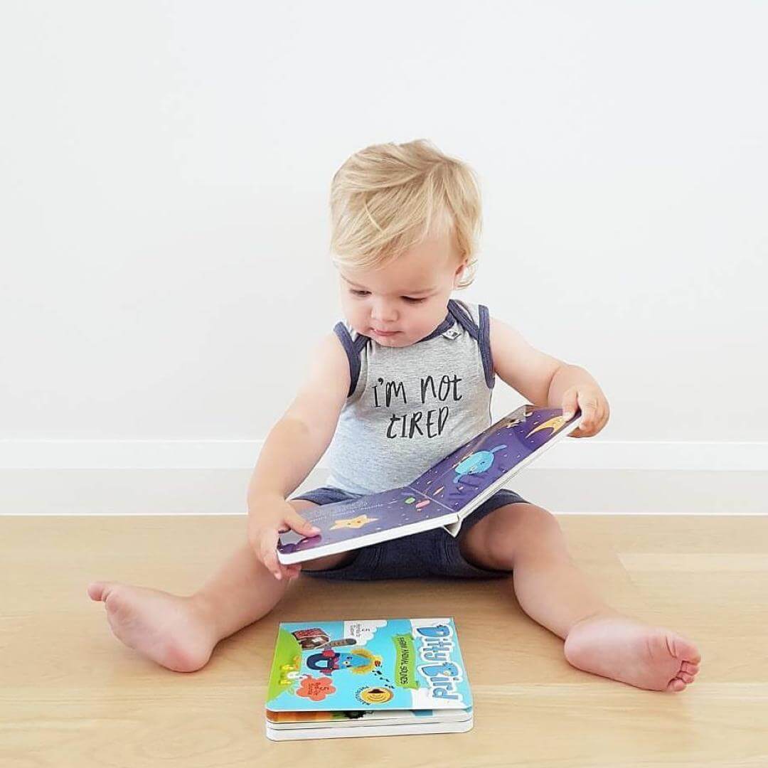 A blonde toddler sits on a wooden floor in a grey sleeveless "IM NOT TIRED" outfit, looking down and holding the Ditty Bird Farm Animal Sounds book. Another colorful book lies nearby.