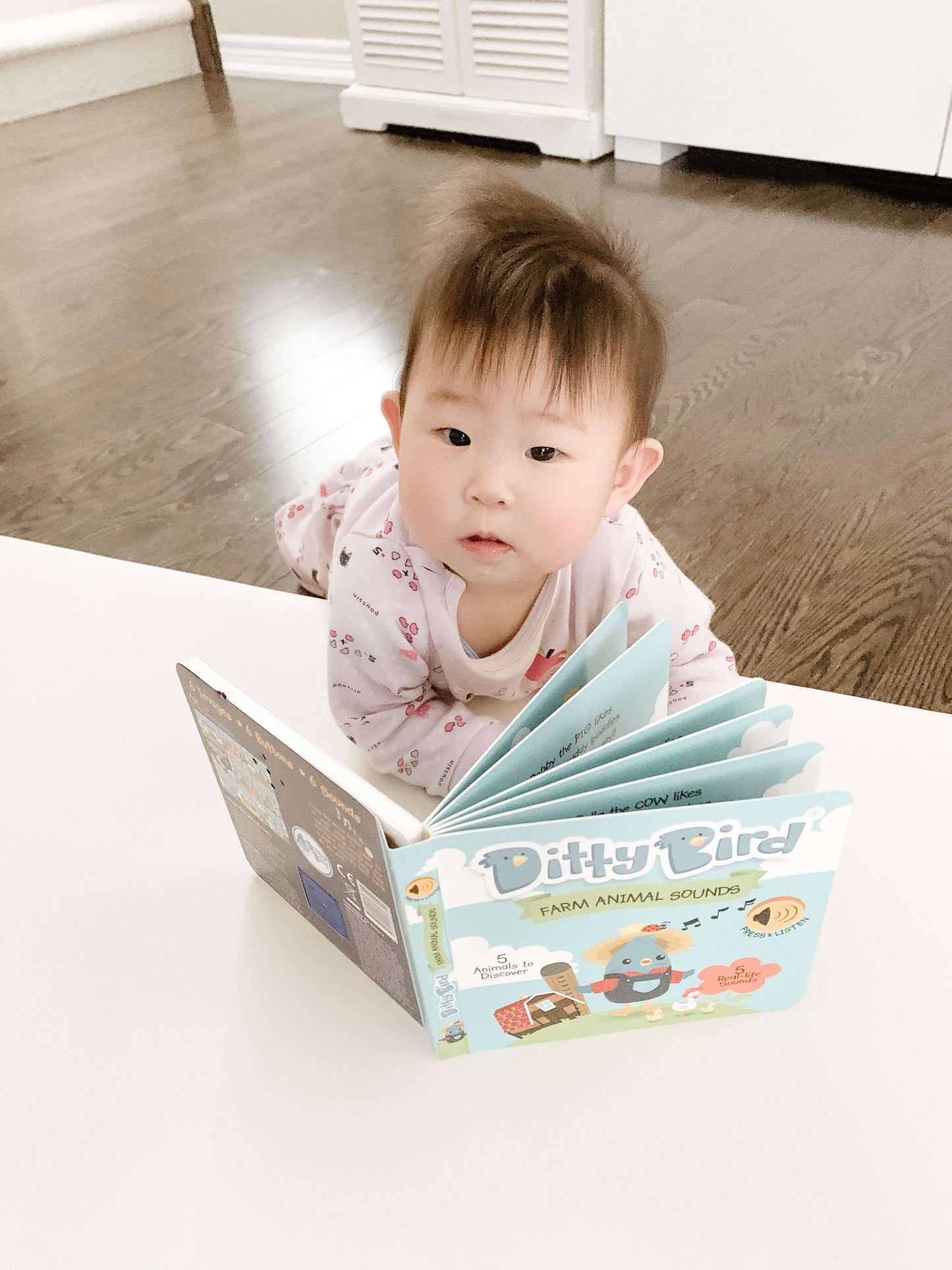 A baby in pajamas lies on their stomach, looking up while holding open the Ditty Bird Farm Animal Sounds book on a white table, with hardwood floors and furniture in the background.