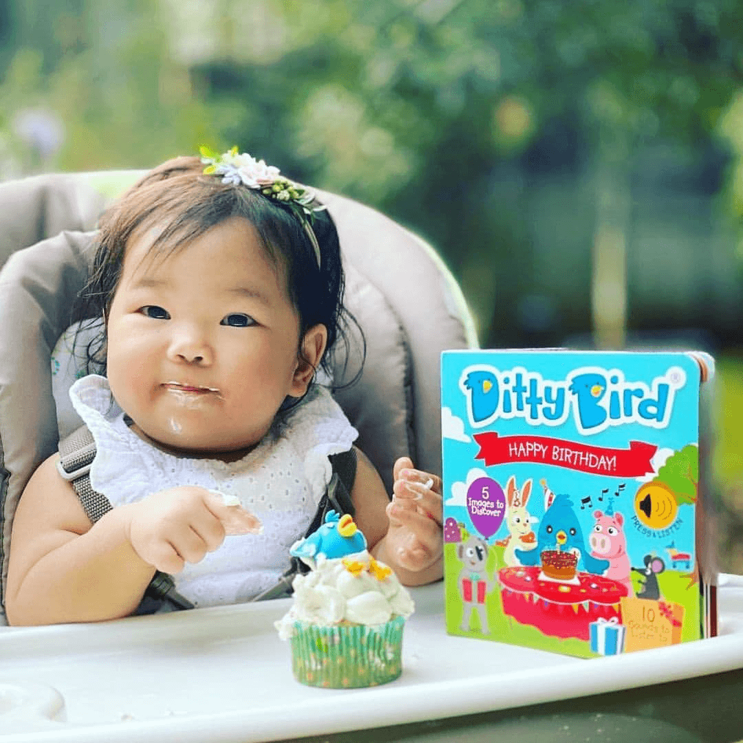 A smiling baby with cake on her face sits in a highchair, holding a cupcake with a bird decoration. Beside her is "Happy Birthday" by Ditty Bird—a colorful musical book and perfect birthday gift for kids. Greenery blurs in the background.