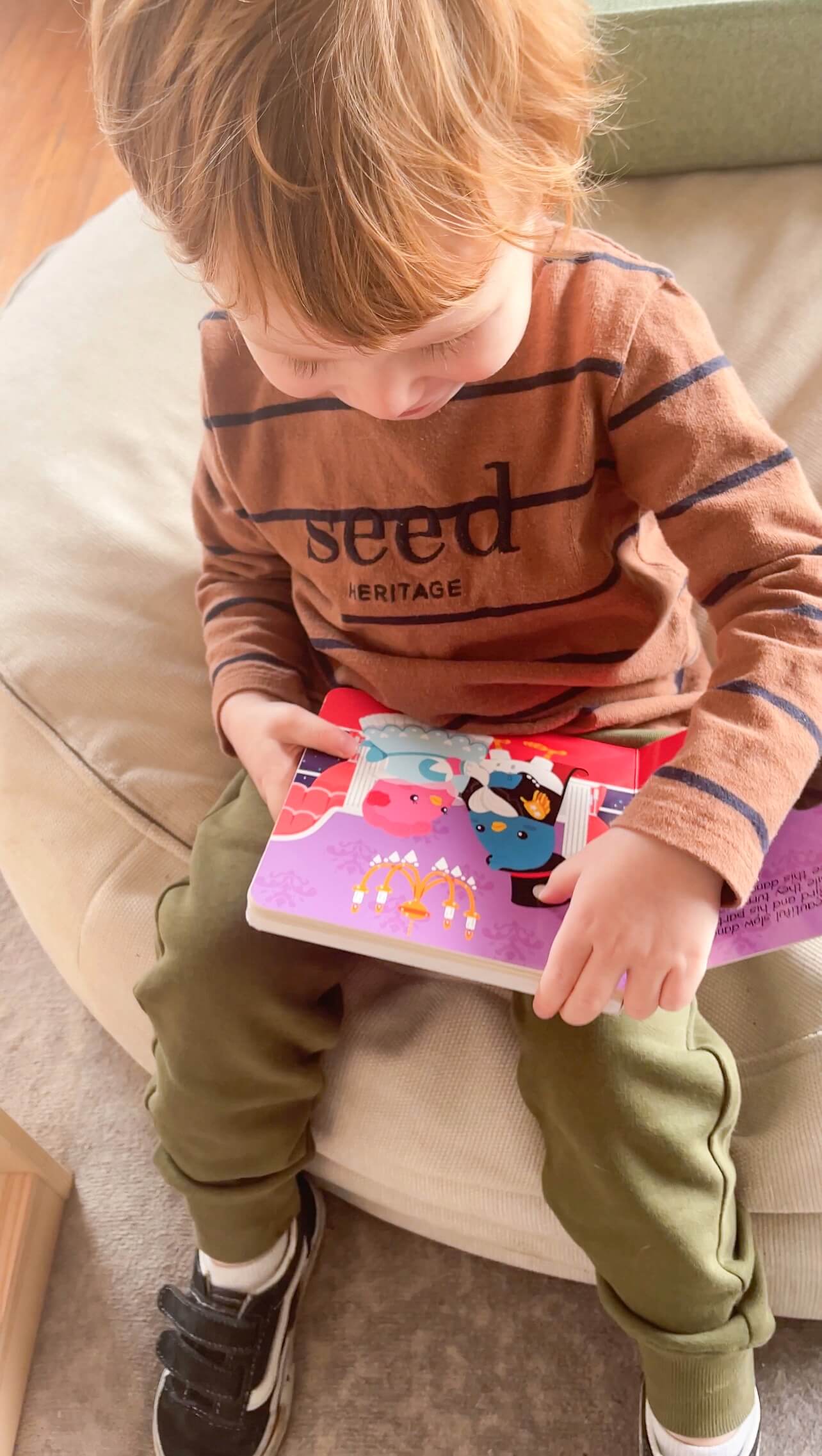 A young child in a brown striped shirt, olive green pants, and black sneakers sits on a beige chair, engaging with the Ditty Bird "Music to Dance to" musical book and touching its colorful pages.