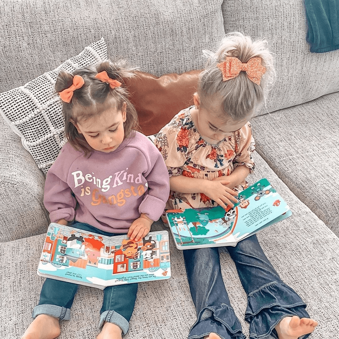 Two young girls sit on a gray couch reading Ditty Bird’s “Music to Dance to,” a musical book with colorful illustrations. One wears a purple “Being Kind is Gangster” sweatshirt; the other wears a floral top and jeans.