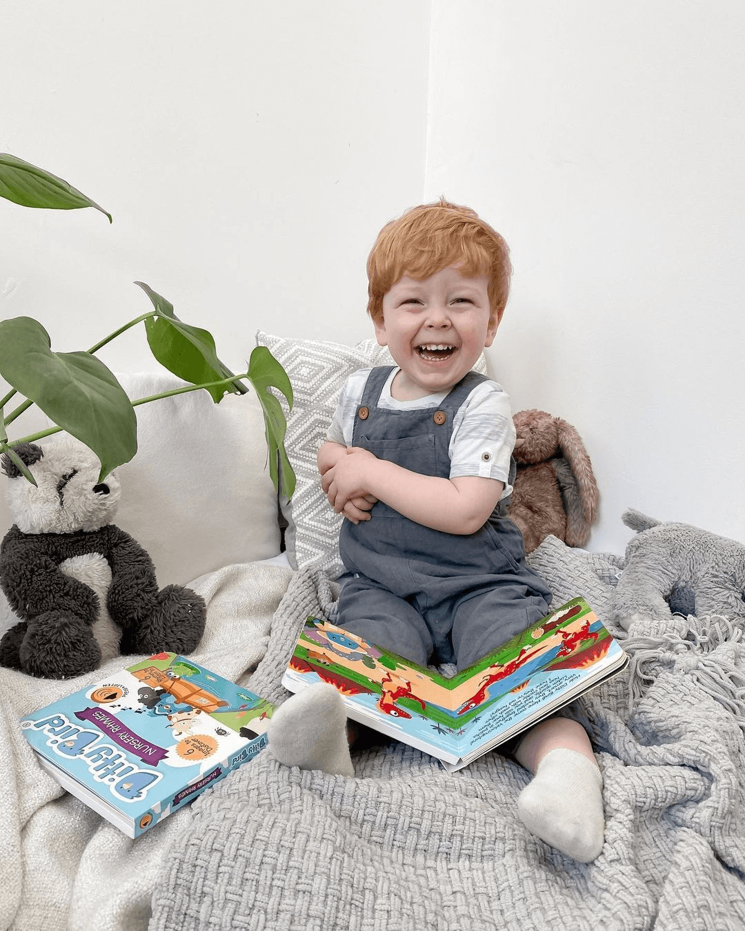 A young child with light red hair smiles happily while sitting on a cozy gray blanket with Ditty Bird’s Dinosaur Sounds book in their lap. Stuffed animals, a plant, and another book are nearby in the bright, cheerful setting.