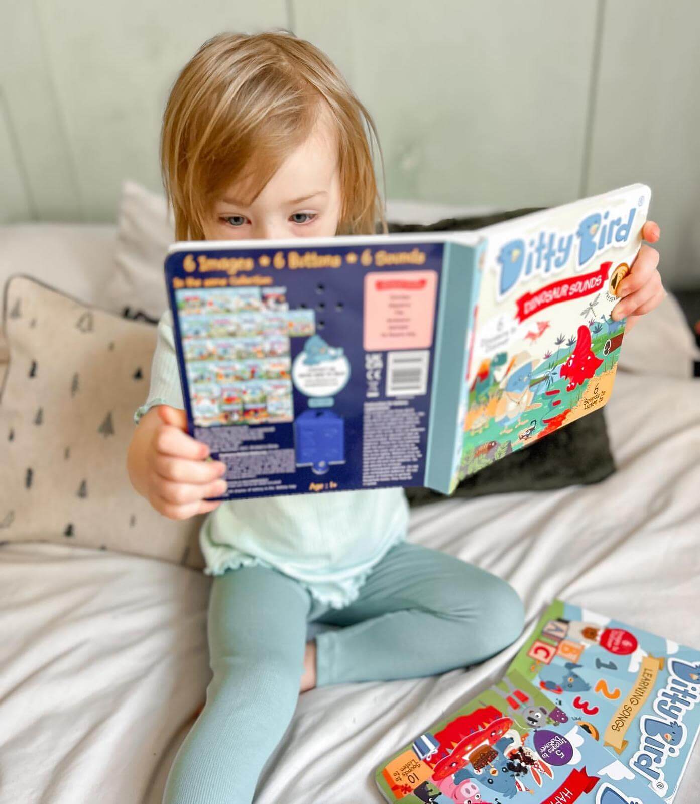 A young child sits on a bed, focused and captivated as they read Ditty Bird’s Dinosaur Sounds book. Another colorful book lies nearby while the fun dinosaur sounds from this interactive Ditty Bird book keep the child engaged.