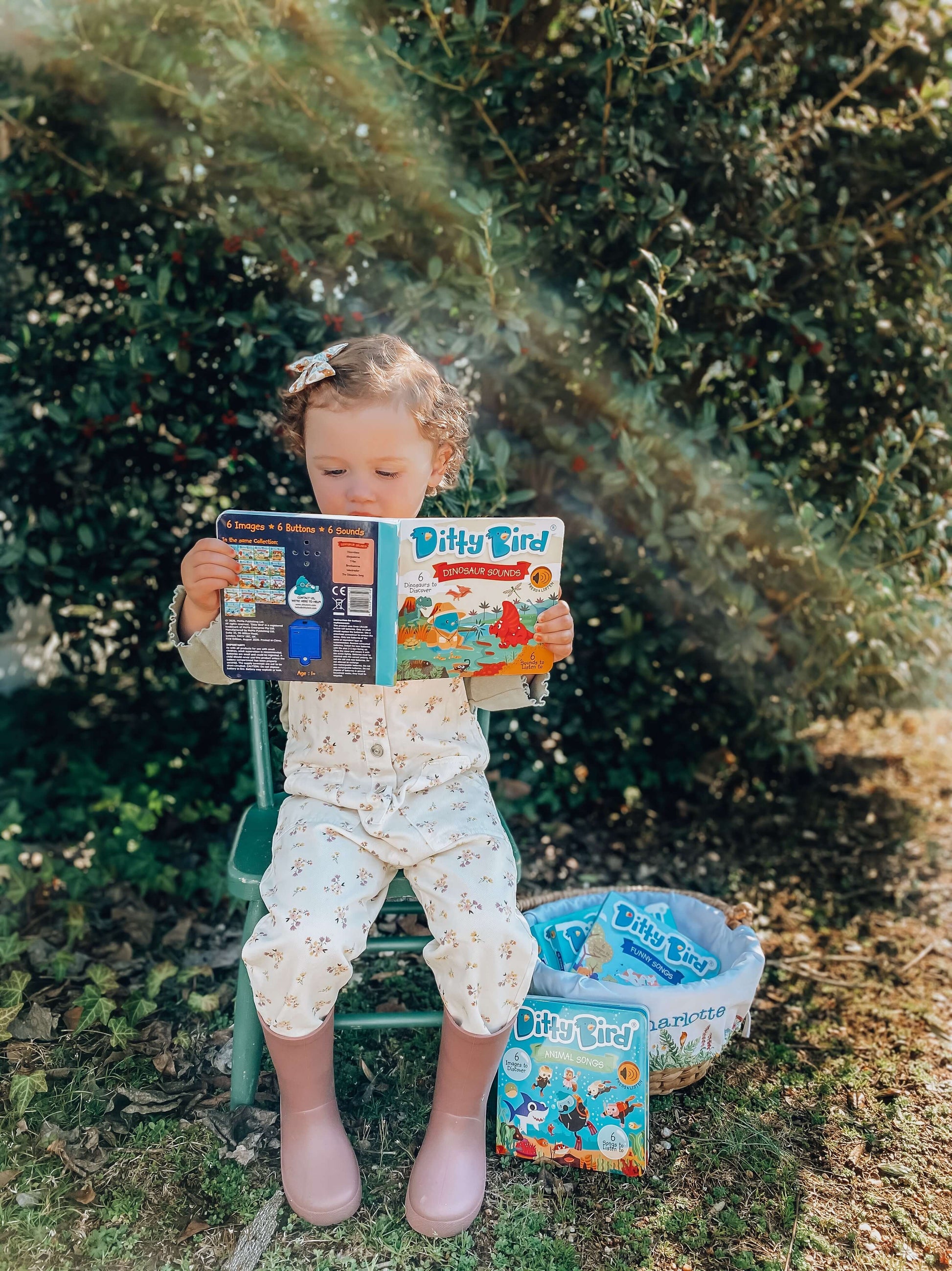 A young child in light floral pajamas and pink boots reads Ditty Bird’s Dinosaur Sounds book outdoors. Next to the chair is a basket filled with more Ditty Bird books. Bushes and greenery can be seen in the background.