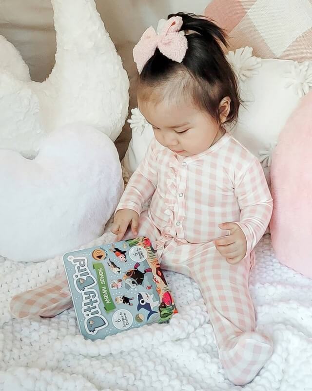 A young child in pink plaid pajamas with a pink bow sits on a white blanket, surrounded by plush pillows and soft toys, looking at the Ditty Bird "Animal Songs" interactive musical book.