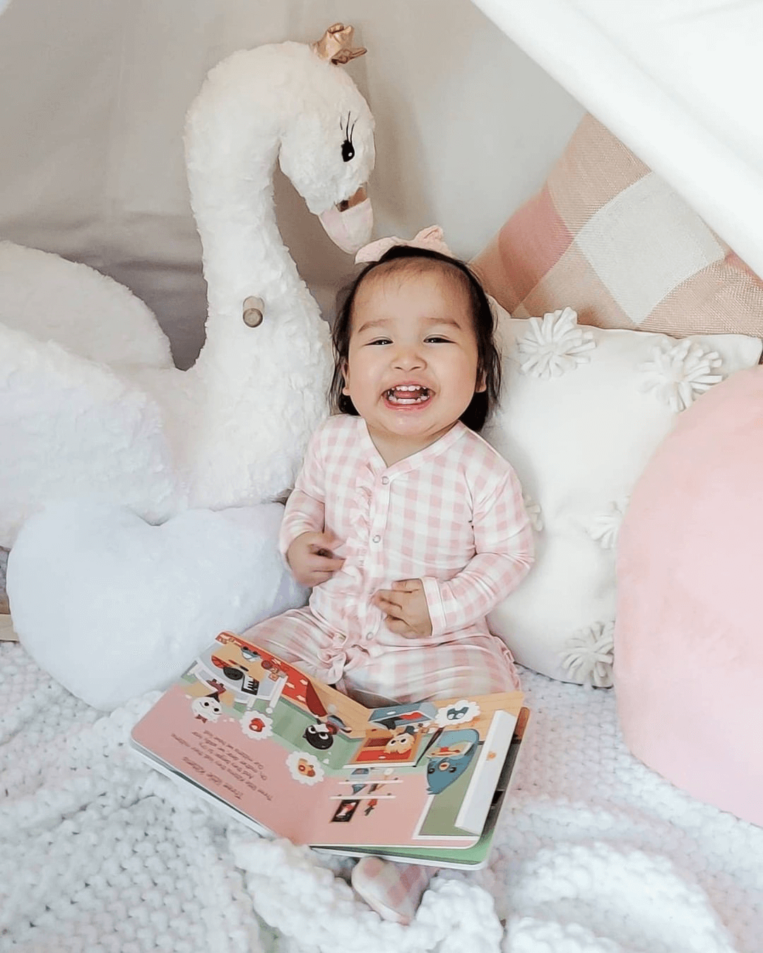 A smiling toddler in pink checkered pajamas sits in a cozy fort with plush pillows, a white stuffed swan, and an open Ditty Bird Animal Songs musical book on their lap. The bright scene radiates cheerfulness.