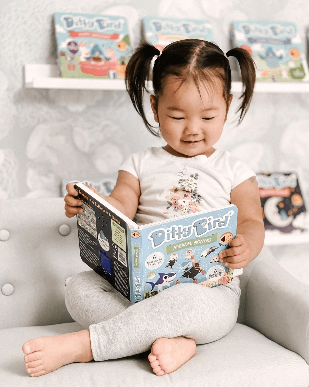 A young girl with pigtails sits cross-legged on a chair, smiling as she reads the Ditty Bird Animal Songs interactive musical book. Children's books can be seen displayed on a shelf in the background.