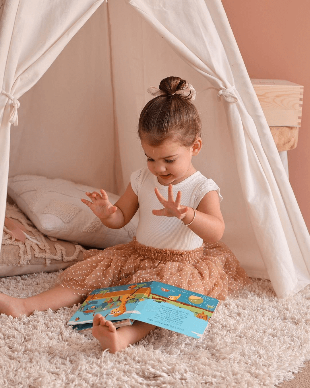 A young girl in a white top and brown tulle skirt sits on a fluffy rug inside a cozy white tent, smiling as she enjoys animal tunes from the Ditty Bird Animal Songs interactive musical book in her lap.