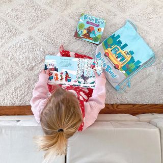 A young blonde-haired child sits on the floor, reading a colorful book. Nearby are Ditty Bird’s Classical Ballet Music book and a blue drawstring bag featuring a car and city illustration.