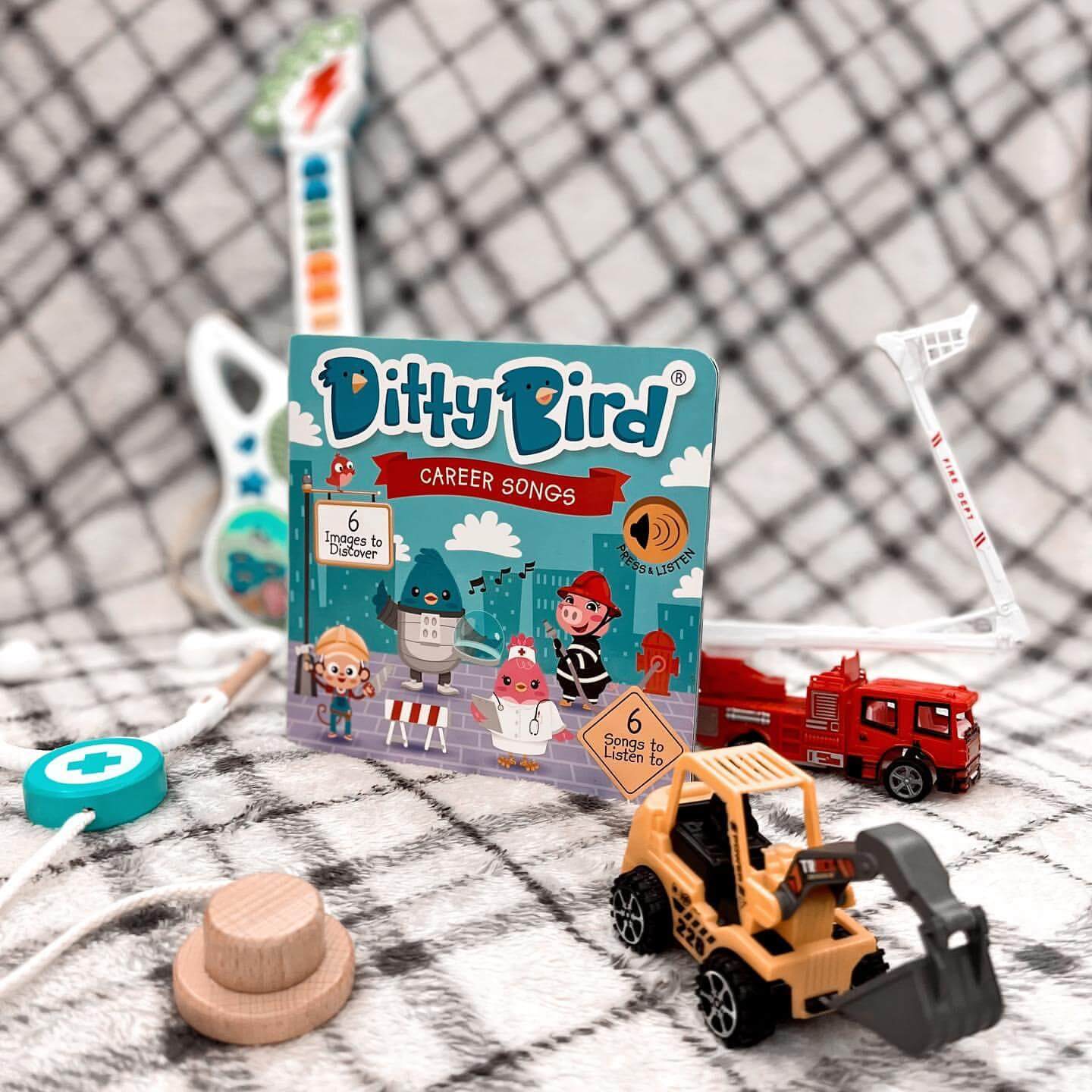Ditty Bird's Career Songs interactive book is displayed with a fire truck, construction loader, toy guitar, stethoscope, and wooden toy on a plaid blanket—a perfect scene for curious preschoolers.