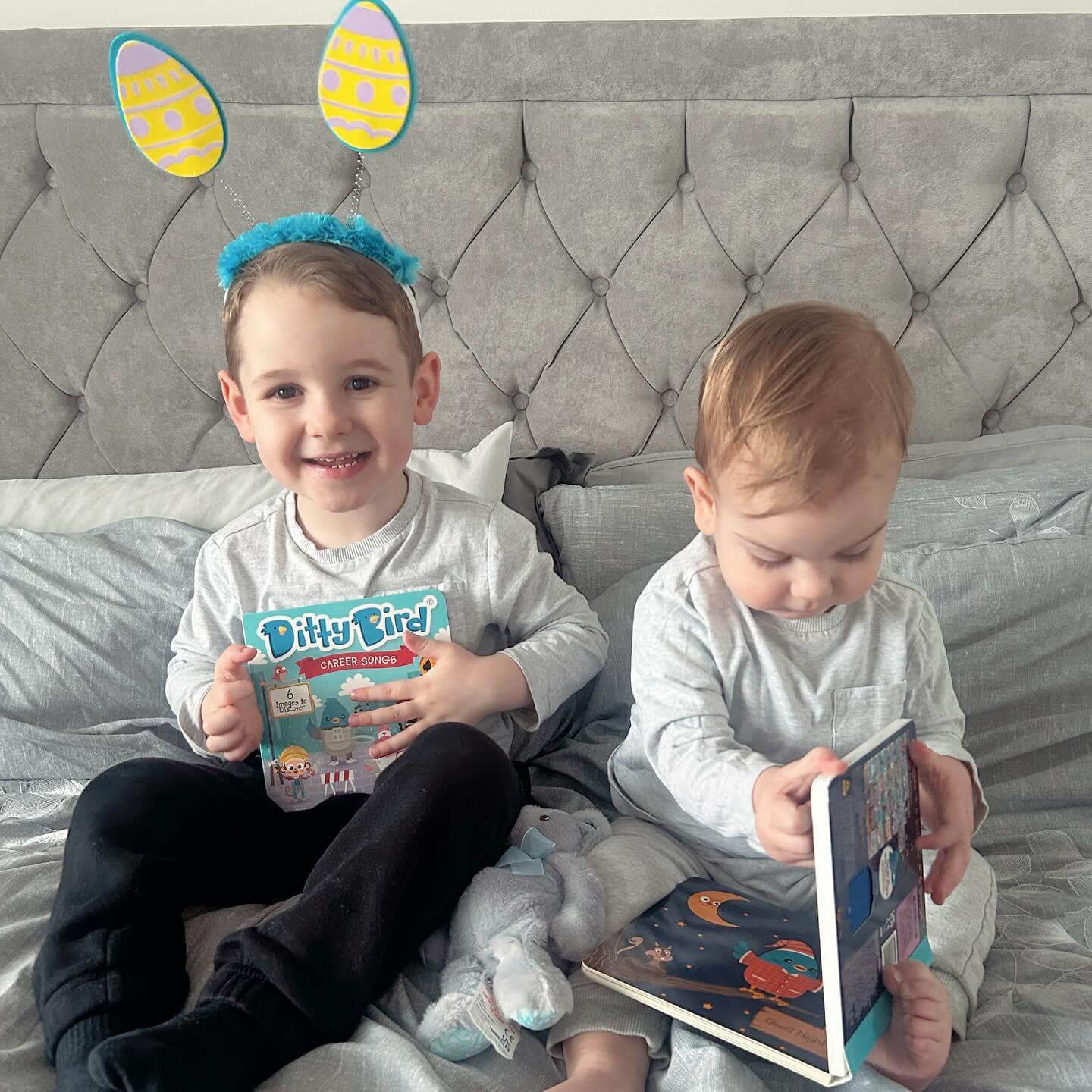 Two preschoolers in matching grey outfits sit on a bed with grey bedding. The older child, wearing blue Easter bunny ears, smiles while holding Ditty Bird’s interactive book “Career Songs.” The younger one looks down, focused on another book.