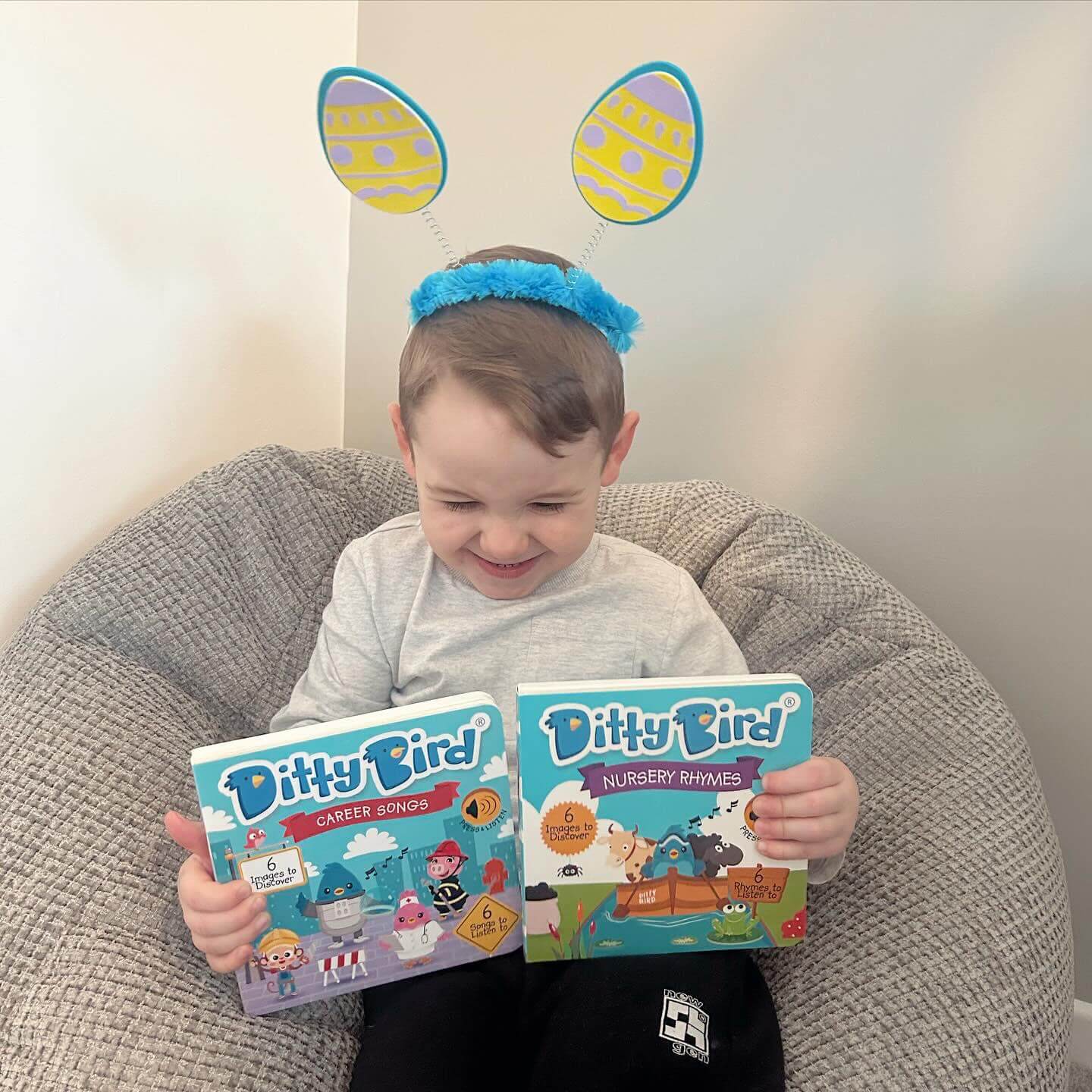 A young child with blue and yellow bunny ear headbands sits on a bean bag, smiling and holding the Ditty Bird "Career Songs" interactive book for preschoolers.