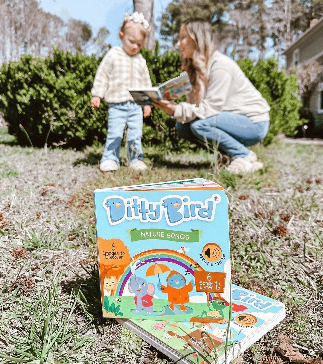 A Ditty Bird "Nature Songs" sound book rests on the grass outdoors, while a woman and young child read together by green bushes on a sunny day.