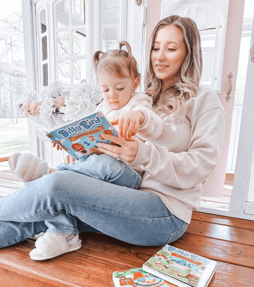 A woman sits on wooden steps with a young child on her lap, reading Ditty Bird’s Nature Songs together. Both wear cozy, light-colored clothes, with more Ditty Bird children’s books scattered nearby.