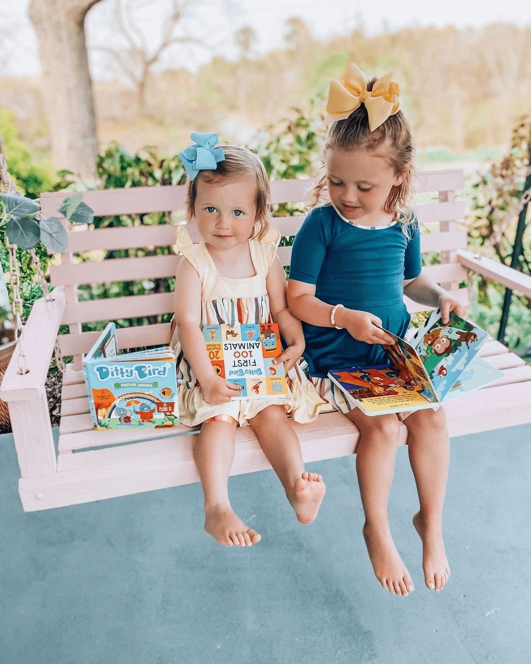 Two young girls with bows in their hair sit barefoot on a pink porch swing, each holding a Ditty Bird "Nature Songs" sound book and smiling. Lush trees and greenery fill the peaceful background.