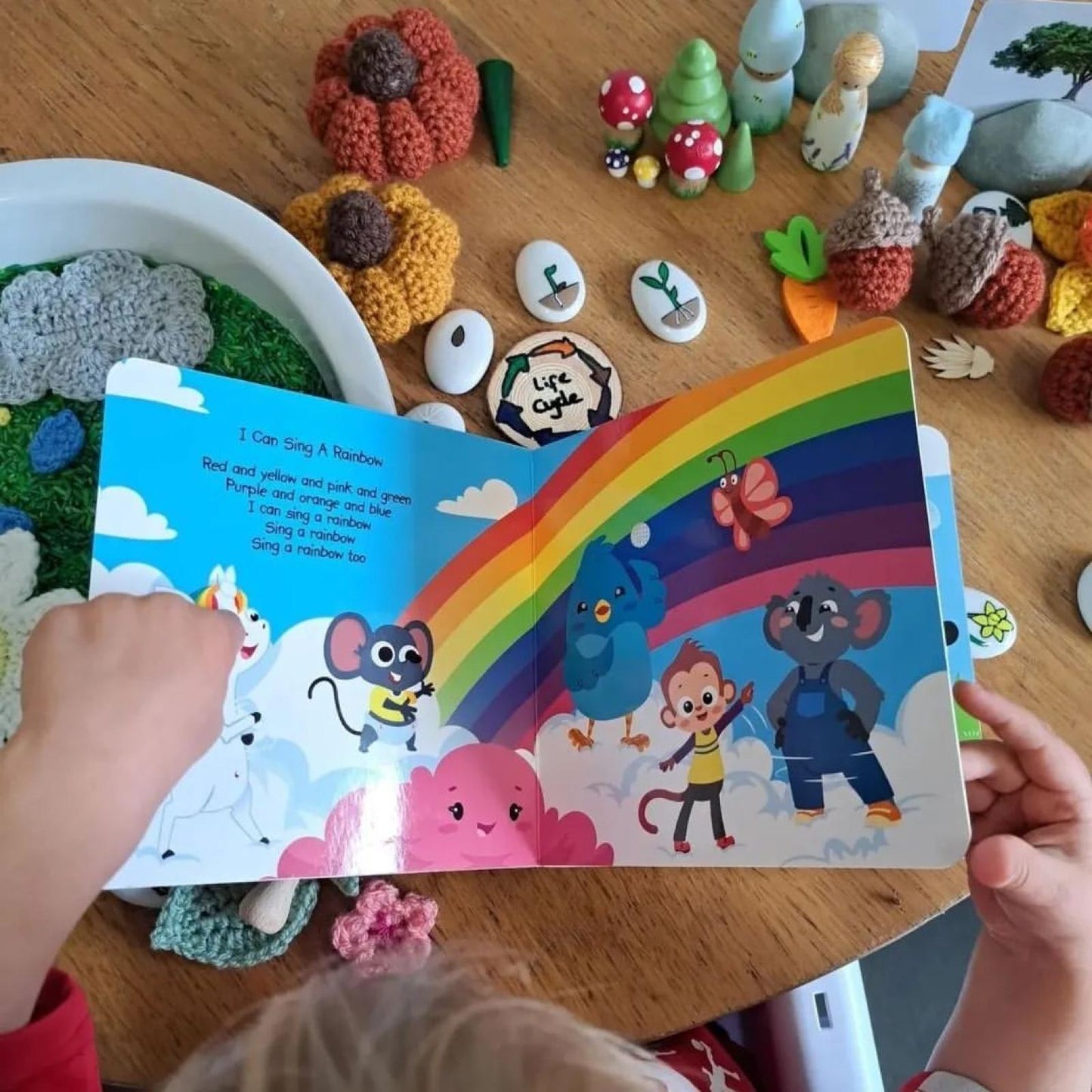 A child reads Ditty Bird’s "Nature Songs," a colorful sound book with cartoon animals under a rainbow. On the wooden table are painted stones, crocheted mushrooms, and toys—creating a creative, playful scene.