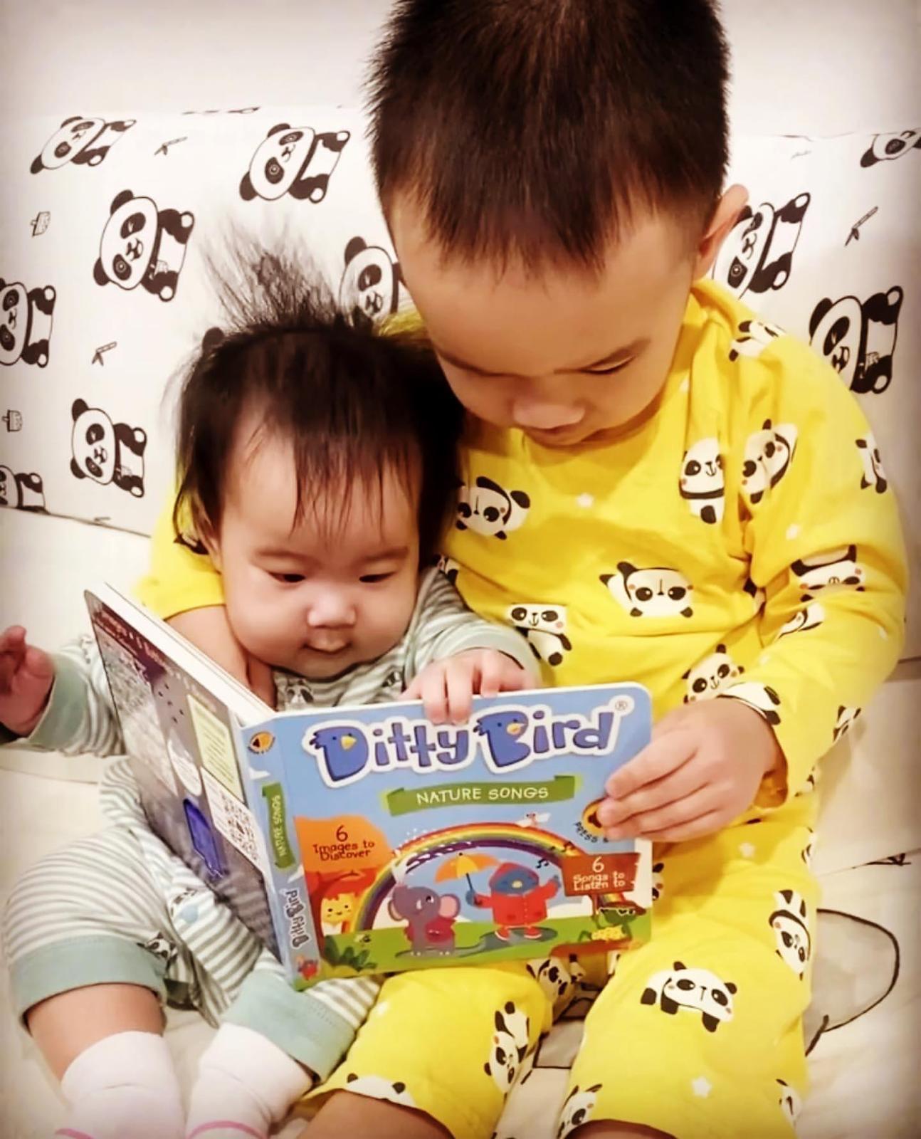 Two young children sit on a panda-print couch, reading the Ditty Bird Nature Songs book. The older child wears yellow panda pajamas, while the baby is dressed in green striped pajamas.