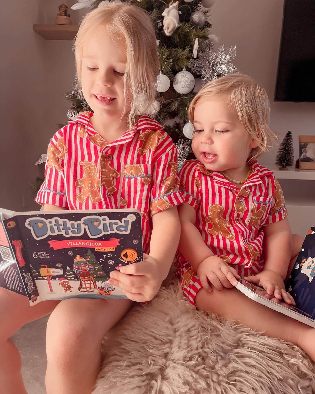 Two young children in matching red and white striped pajamas sit by a decorated Christmas tree, smiling as they read Ditty Bird's "Villancicos en Español," a sound book filled with Spanish Christmas songs.