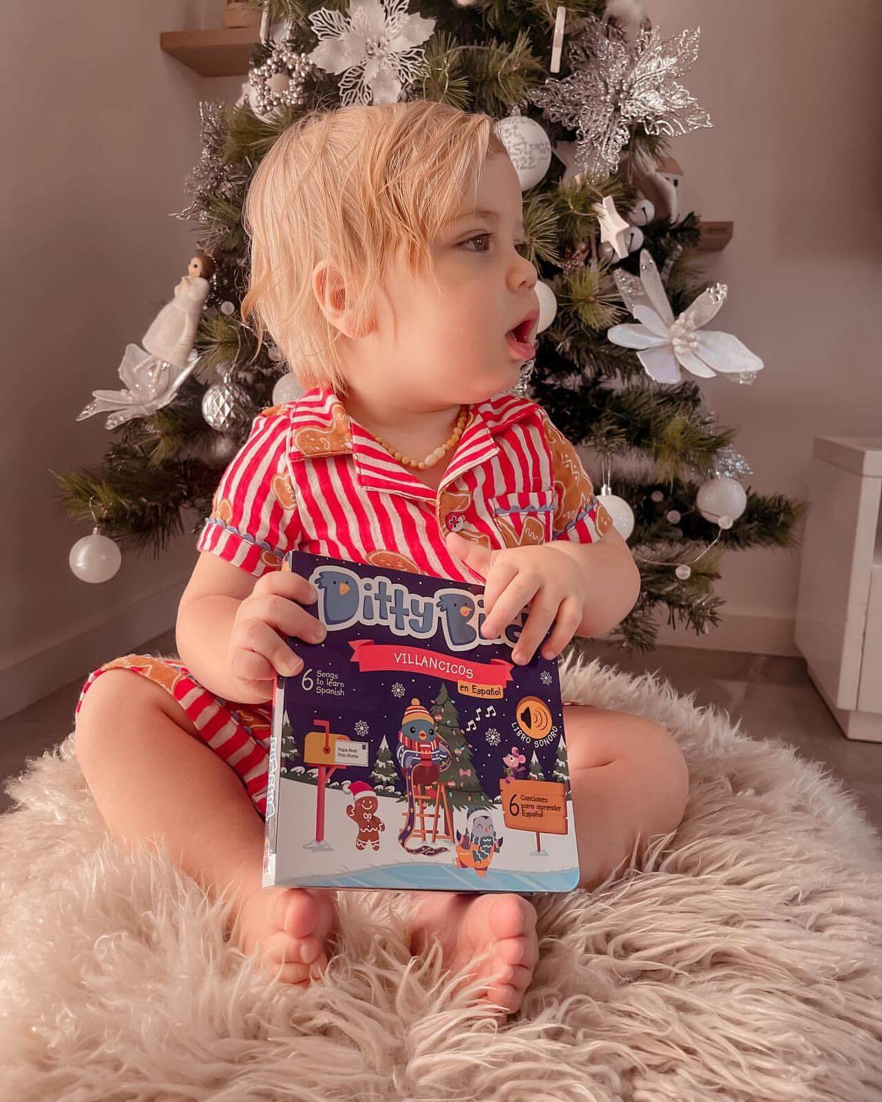 A young child in red-striped pajamas sits on a fluffy white rug by a decorated Christmas tree, holding the Ditty Bird Villancicos en Español sound book and looking to the side.