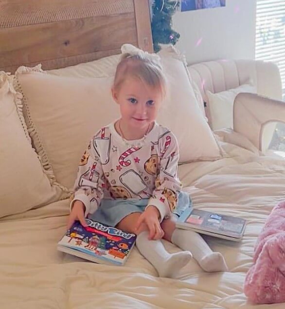 A young child with a light bow sits on a neatly made bed, holding Ditty Bird’s "Villancicos en Español" sound book and a closed laptop. She wears a white sweater with playful designs, blue skirt, and knee-high socks.