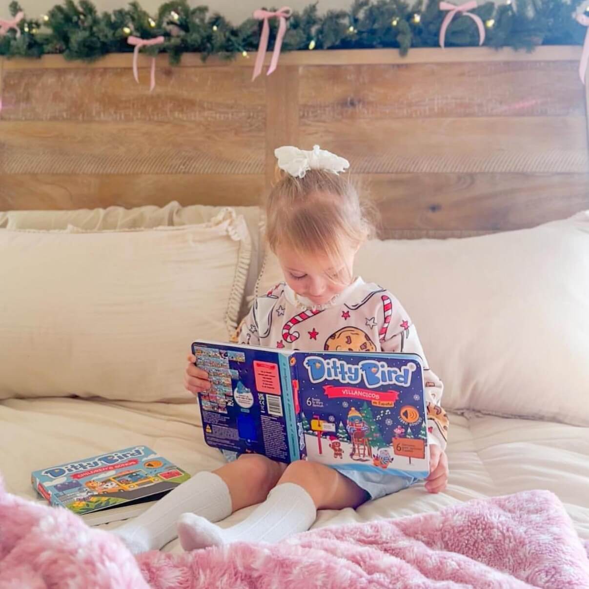 A young girl with a white bow sits on a bed, wearing pajamas and reading the Ditty Bird "Villancicos en Español" sound book. She is surrounded by pillows, a pink blanket, another book, and holiday garland above the headboard.