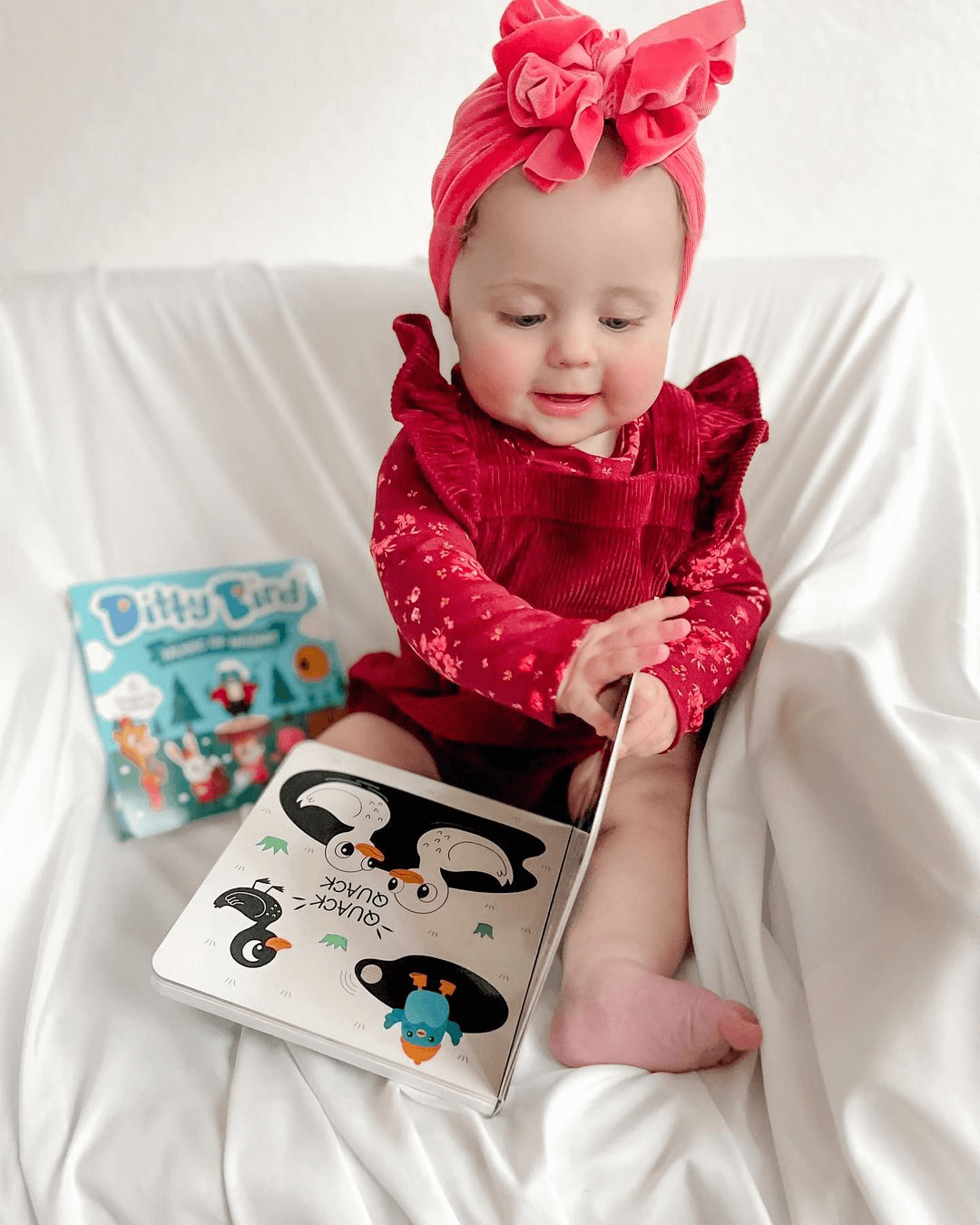 A baby in a red outfit and headband sits on a white blanket, smiling as she turns the pages of Ditty Bird's Black & White Animals high-contrast sound book. Another colorful children’s book lies beside her.