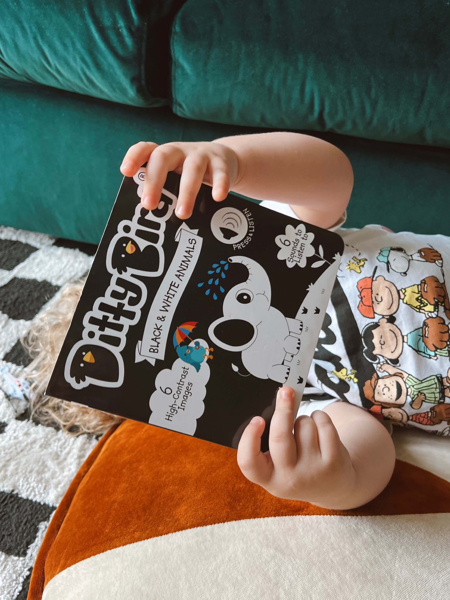 A toddler lounges on a patterned rug by a green sofa, holding Ditty Bird's "Black & White Animals" book, wearing a cartoon character shirt and enjoying the charming high-contrast animal illustrations on the cover.