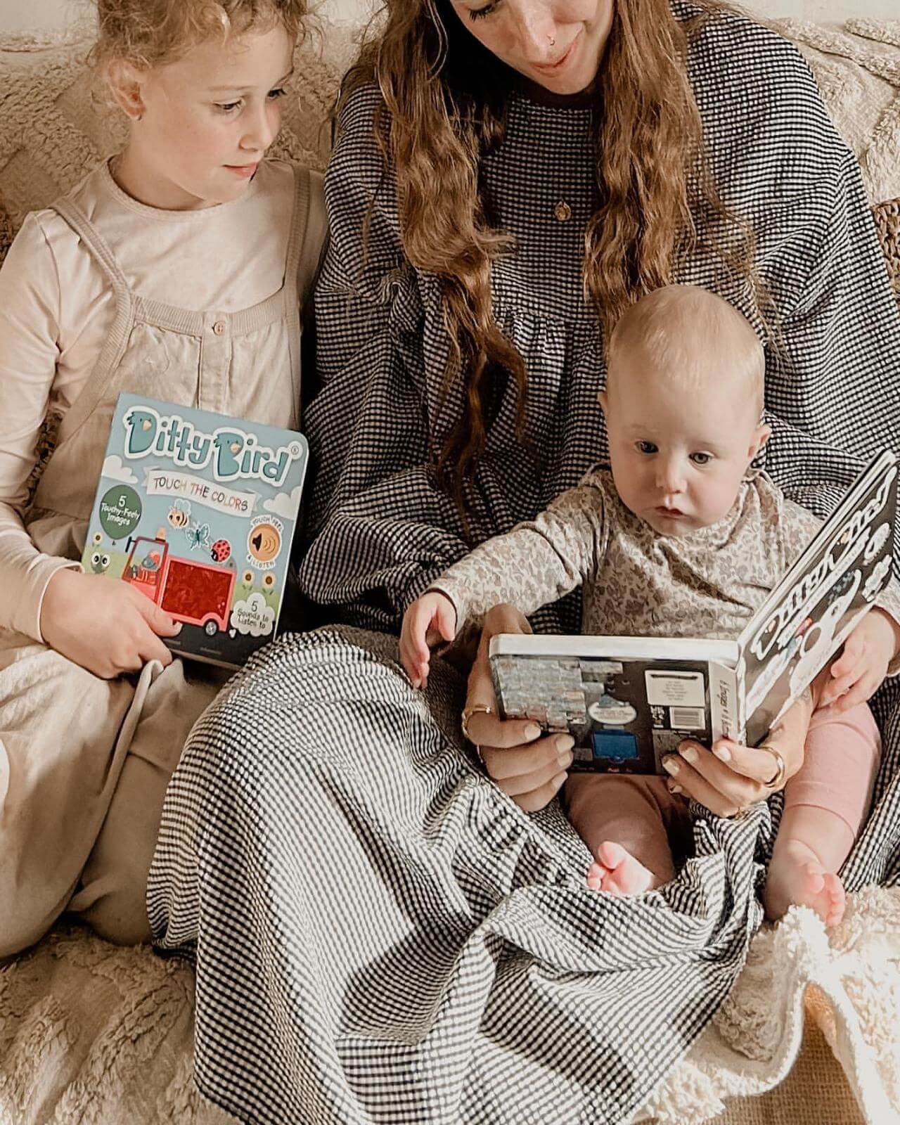 A woman sits on a couch with a baby on her lap and a young child beside her. The baby looks at Ditty Bird's Black & White Animals book, while the child smiles, holding another colorful Ditty Bird book.