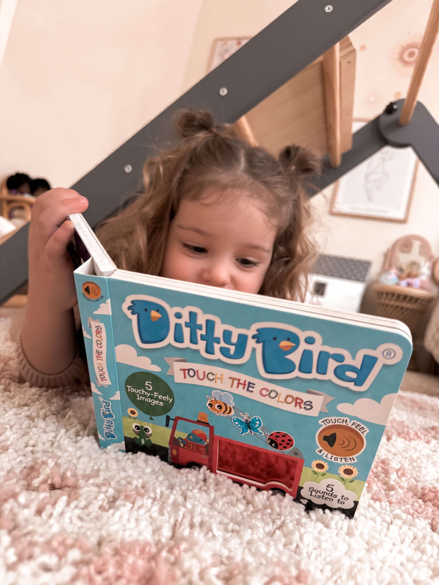 A young child with brown pigtails lies on a patterned rug under a wooden play structure, exploring Ditty Bird's Touch the Colors | USB-C rechargeable sound book and enjoying its vibrant cover illustrations.