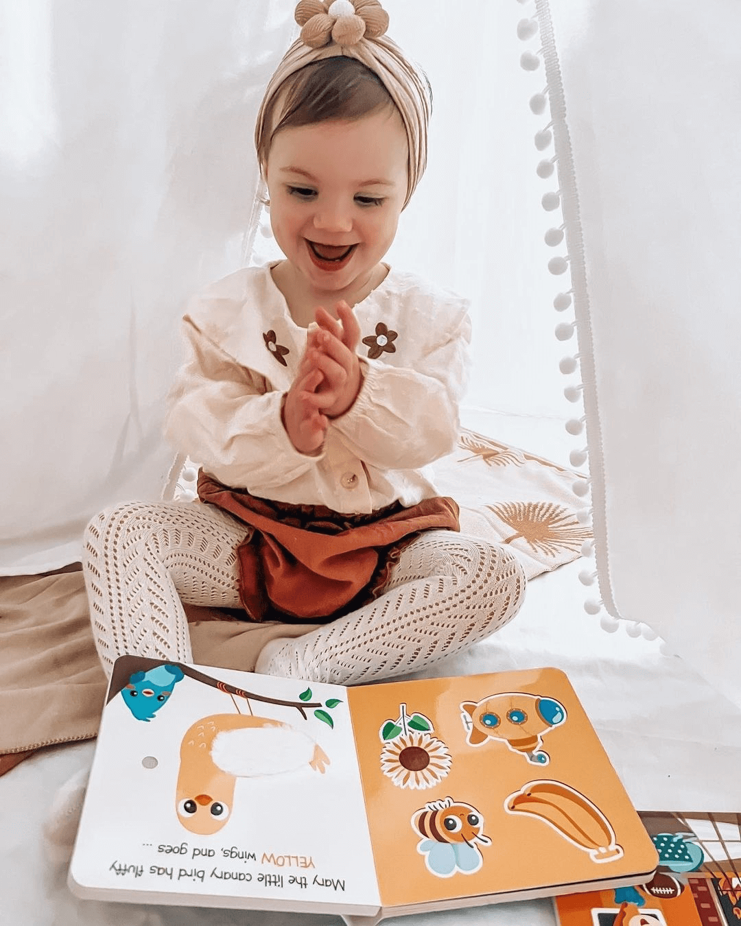 A smiling young child sits cross-legged, clapping joyfully while exploring the Ditty Bird Touch the Colors sensory book (USB-C rechargeable) spread open in front of her inside a cozy tent.