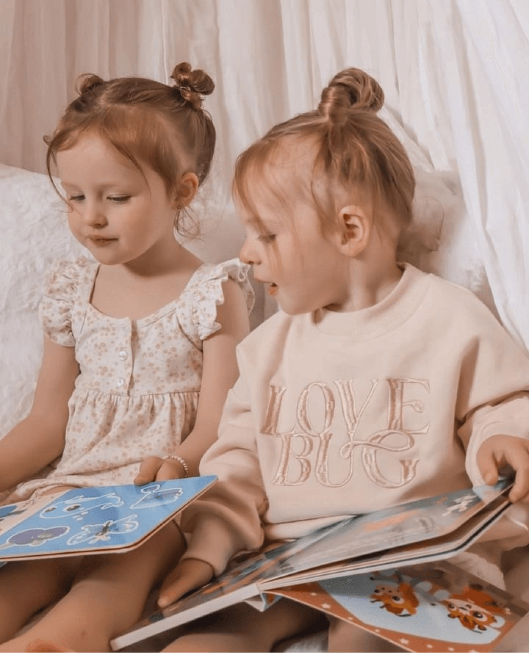 Two young girls with light brown hair in small buns sit on a white bed, exploring Ditty Bird's Touch the Colors sensory book (USB-C rechargeable). One wears a floral dress, the other a pale sweatshirt with "LOVE BUG" on it.
