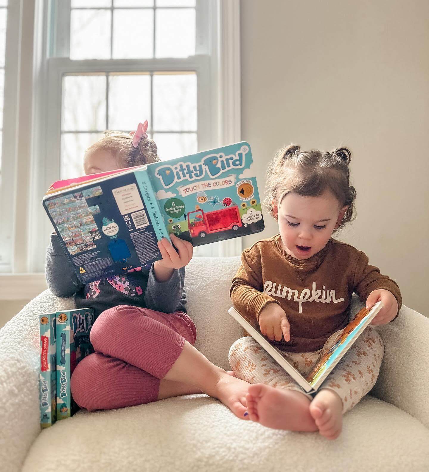Two young children sit together by a window, each reading. One holds up Ditty Bird’s Touch the Colors, a USB-C rechargeable sound book, while the other points excitedly at a page. Both look focused and immersed in their books.