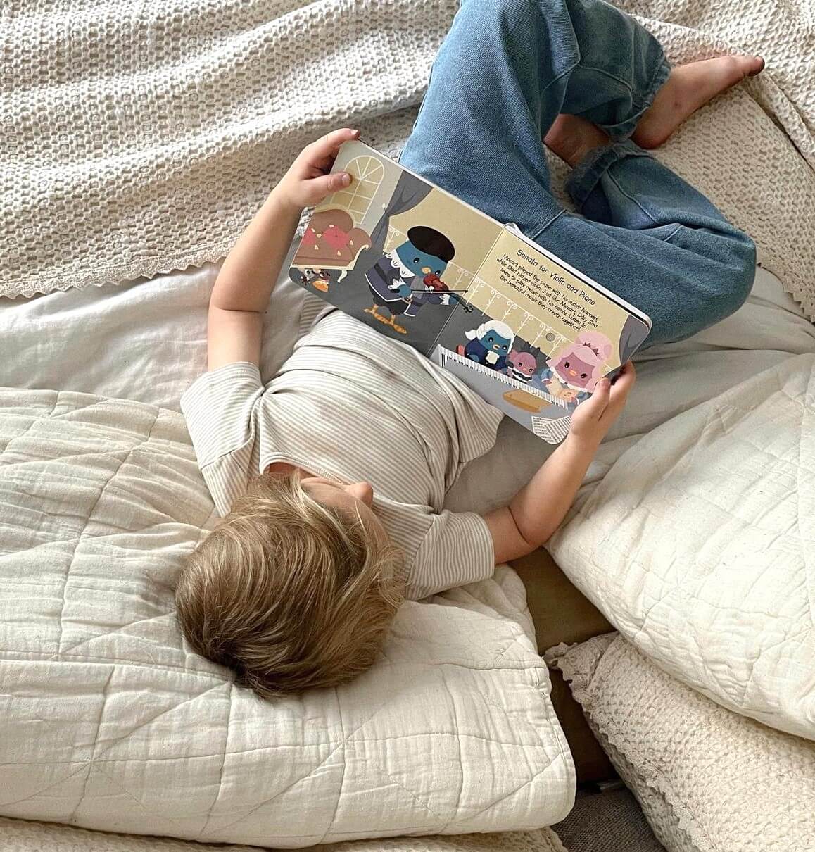 A young child lies on a bed with white bedding, wearing a striped shirt and blue pants, reading Ditty Bird's "Music of Mozart," an interactive children's book.