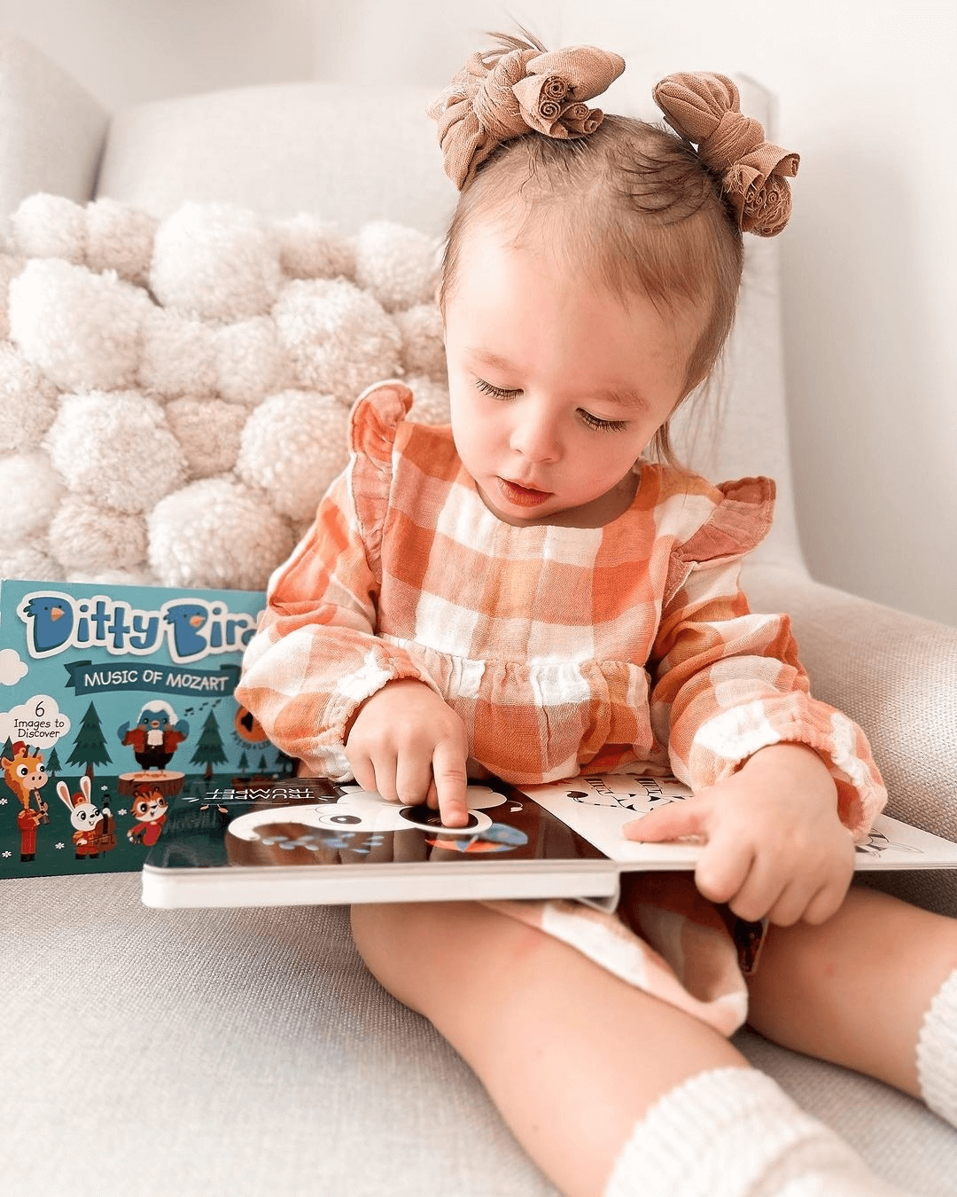 A young girl with two hair buns in a peach plaid dress sits in a chair, pointing at the pages of an interactive children’s book. Next to her is the Ditty Bird Music of Mozart book for kids.