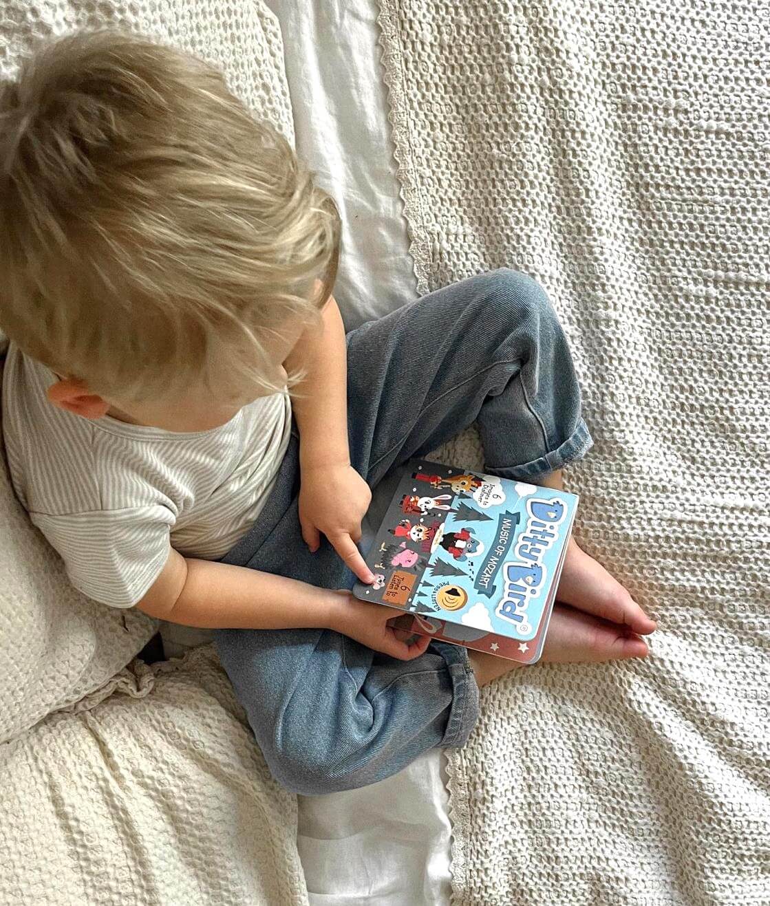 A young child with blonde hair sits on a bed with cream bedding, wearing a striped shirt and blue pants, pointing at the page of Ditty Bird's "Music of Mozart," an interactive children's book featuring classical music for kids.